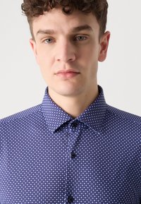 Camisa azul con un patrón, con cuello tipo polo, que presenta pequeñas formas geométricas. La textura parece suave, con botones negros y un cuello ajustado.