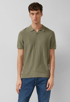 Junger Mann mit lockigem Haar, der ein olivgrünes kurzärmeliges Poloshirt und blaue Jeans trägt, steht vor einem hellgrauen Hintergrund.