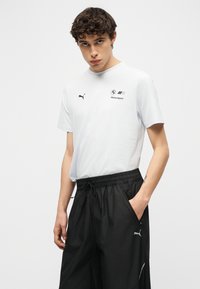 Νεαρός άνδρας που φοράει λευκό μπλουζάκι BMW Motorsport Puma και μαύρο παντελόνι φόρμας Puma, με το ένα χέρι στην τσέπη, μπροστά σε απλό λευκό φόντο.