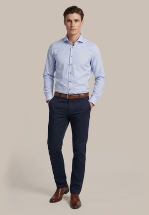 Man staat met handen in de zakken, draagt een lichtblauw overhemd, donkerblauwe broek, bruine riem en bruine nette schoenen op een beige achtergrond.