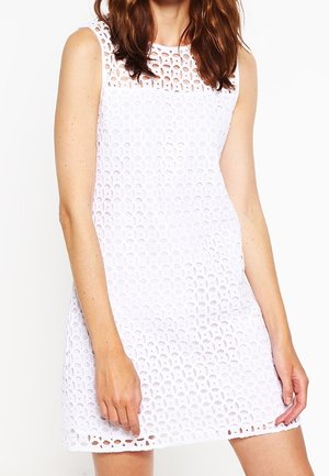 Frau trägt ärmelloses weißes Kleid mit Häkelmuster, steht mit einer entspannten Hand und der anderen leicht angewinkelt.