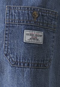 Denim fabric in deep blue with a visible stitched pocket, featuring a white label that reads "GUESS JEANS" with a branded button above.
