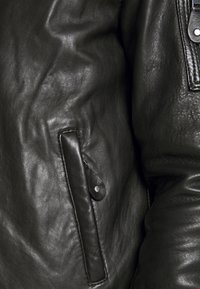 Schwarze Lederjacke mit sichtbarem Reißverschluss, strukturiertem Muster und einer Außentasche mit Druckknopfverschluss. Weiche, glatte Verarbeitung.