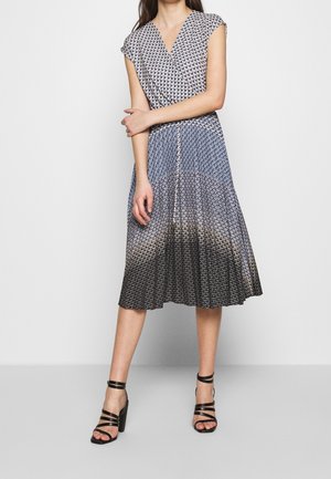 Femme portant une robe midi plissée à manches courtes et col en V, avec un motif en écailles noir, blanc et bleu, assortie à des talons noirs à brides.