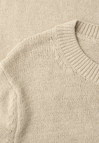 Gros plan sur le tissu en tricot beige d'un pull montrant un col ras du cou côtelé et un motif de points texturés.