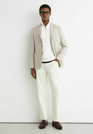 Mees, kes kannab musti prille, beežikat ruudulist pintsakut, valget lukuga kampsunit, kreemikarva pükse ja pruune madalaid jalanõusid, seisab valge seina ees.