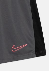 Harmaa urheilujersey, jossa on mustat verkkokohdat ja näyttävä punainen brodeerattu Nike-logo alhaalla edessä. Silkkinen pinta, moderni muotoilu.