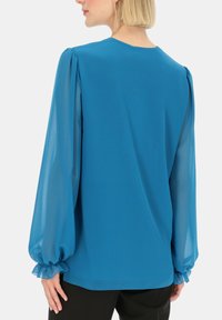 Niebieska bluzka z gładkiego materiału i przezroczystymi bufkami. Posiada elastyczne mankiety i klasyczny okrągły dekolt. Minimalistyczny design.