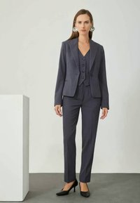 Dresser i mørkegrått tre-delt sett som består av en tilpasset blazer, matchende vest med knapper og skreddersydde bukser, komplementert med svarte hæler.