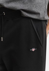 Primo piano di pantaloni da ginnastica neri con tasche anteriori, vita con coulisse e piccolo logo GANT rosso, bianco e blu sulla coscia, indossati con un top grigio.