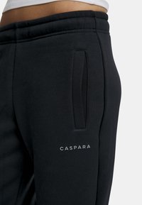 Sorte joggebukser med en glatt tekstur, elastisk midje, sidelommer og en "CASPARA"-logo på venstre side.