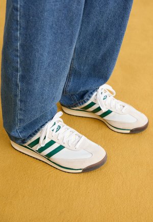 Hvide sneakers med grønne striber båret med blå jeans på en sennepsfarvet gulv.