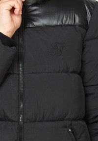 Svart pufferjacka med ett glänsande svart nylontyg i övre delen, dragkedjestängning, ribbade ärmslut och en subtil broderad logotyp på bröstet.