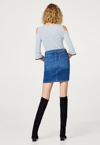 Falda de mezclilla con forma ajustada, color azul y dos bolsillos traseros. Combina con una blusa gris claro con cortes al hombro y mangas acampanadas.