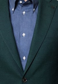 Zielony blazer o fakturze z jednym guzikiem, noszony na niebieskiej koszuli w wzór jodełki z białymi guzikami i spiczastym kołnierzykiem.