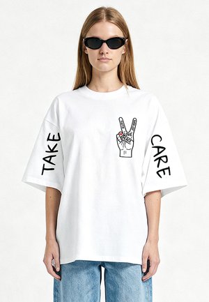 Femme aux longs cheveux portant des lunettes de soleil noires et un t-shirt blanc oversize avec "TAKE" sur la manche gauche, "CARE" sur la droite, et un motif de signe de paix avec la main.