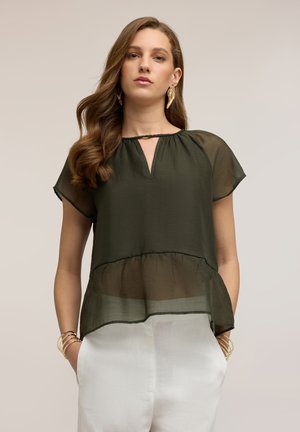 Donna con capelli castani ondulati che indossa una camicetta verde oliva trasparente con scollo a goccia e pantaloni bianchi, mani nelle tasche, sfondo neutro.