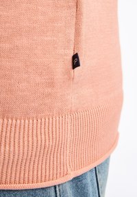 Hellbeige gestrickter Pullover mit geripptem Saum und einem kleinen Stoff-Logosticker in Marineblau. Die Textur wirkt weich und glatt.