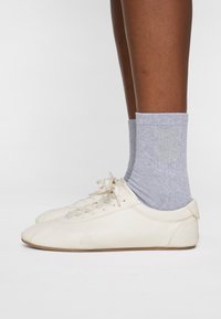Hvide lædersneakers med struktureret overflade, tynde snørebånd og gummisåler, parret med grå ankelstrømper med et skinnende cirkulært mønster.