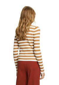 Femme aux cheveux longs et ondulés portant un haut à manches longues côtelé à rayures marron et blanc et un pantalon couleur rouille, vue de dos.