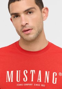 Czerwony bawełnowy T-shirt z okrągłym dekoltem i białym napisem "MUSTANG" z "JEANS COMPANY SINCE 1932" pod spodem.