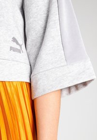 Top de manga corta gris claro de Puma llevado sobre una falda de pliegues naranja, mostrando la parte media del brazo y el torso lateral.