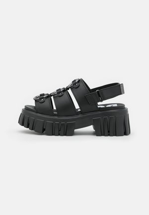 Black platform sandals with multiple straps, featuring metal accents and a chunky, ridged sole for added texture and height.