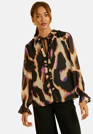Žena nosi blúzku s dlhými rukávmi s abstraktným farebným zvieracím vzorom, s volánikmi vpredu a detailom na viazanie okolo krku, v kombinácii s čiernymi nohavicami.