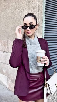 Sorte solbriller, grå strikket turtleneck, maroon blazer og lær miniskjørt. Holder en kaffekopp, med en hvit handveske.