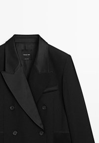 Schwarzer, doppelt geknöpfter Blazer mit Satin-Revers, Pattentaschen und tonaler Naht. Etikett sichtbar im Krageninneren.