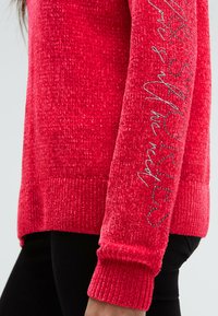 Roter, strukturierten Pulloverärmel mit silberner Schrift und Nieten-Design, getragen über schwarzen Hosen vor einem schlichten Hintergrund.