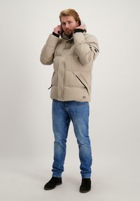 Lichtbeige pufferjack met een capuchon, frontale ritssluiting en twee zijzakken. Draagt bij blauwe jeans en zwarte enkellaarzen. Textuurstofafwerking.