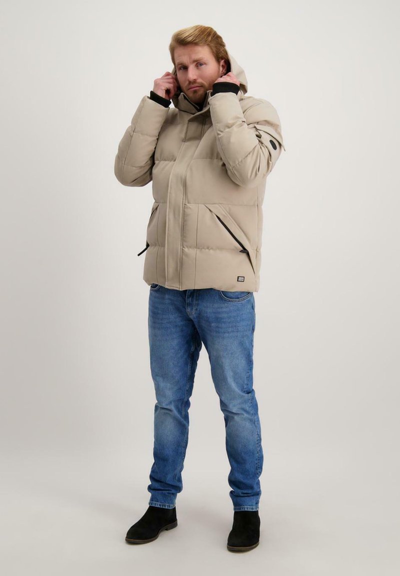 Lichtbeige pufferjack met een capuchon, frontale ritssluiting en twee zijzakken. Draagt bij blauwe jeans en zwarte enkellaarzen. Textuurstofafwerking.