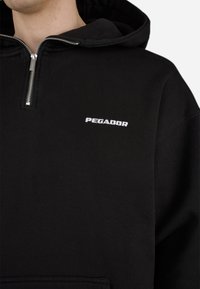Sort hættetrøje med halvlynlås fortil, blødt stof, snøre i hætten og "PEGADOR" logo i hvidt broderet på brystet.