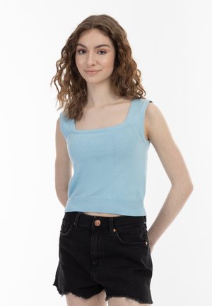 Tânără cu păr castaniu creț, purtând un top bleu fără mâneci și pantaloni scurți din denim negri, stând cu mâinile la spate.
