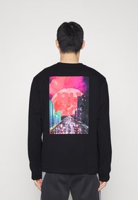 Mann, der sich abwendet, trägt ein schwarzes Langarmshirt mit einem Grafikmotiv von nächtlichem Stadtverkehr, pinkem Himmel und einem großen roten Mond auf dem Rücken.