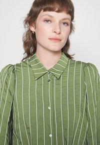 Blusa verde com riscas brancas verticais, abotoada à frente, com colarinho e mangas bufantes; tecido texturizado com uma aparência ligeiramente enrugada.