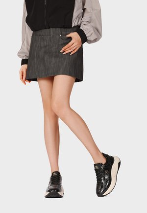 Donna con minigonna nera, giacca nera e grigia e sneakers nere con plateau in piedi con una gamba leggermente piegata.