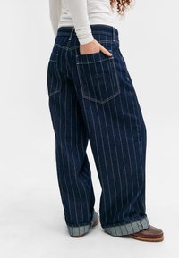 Jean en denim à jambes larges en indigo foncé avec des rayures blanches. Comprend deux poches arrière, des détails cousus et des ourlets retroussés.