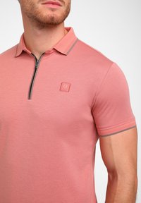 Šviesiai rožinės spalvos trumpų rankovių polo marškiniai su užtrauktuku kaklo srityje, kontrastinės spalvos apykaklės apdaila ir logotipo etikete ant krūtinės. Lygus audinys.