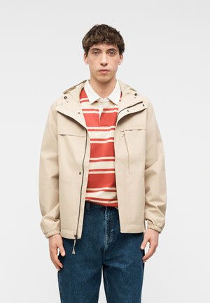 Νέος άνδρας που φοράει μπεζ μπουφάν με κουκούλα πάνω από κόκκινο και άσπρο ριγέ πόλο πουκάμισο και μπλε τζιν, στέκεται μπροστά σε απλό φόντο.