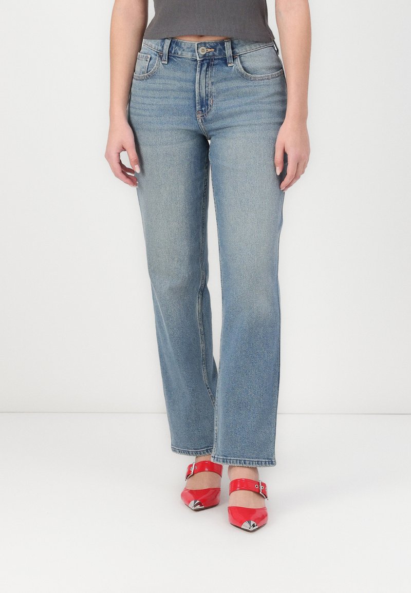 Jeans de ganga azul claro com um design de perna reta, estilo de cinco bolsos e fecho de botão. Usados com sapatos de salto alto vermelhos.