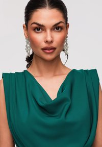 Femme aux cheveux foncés tirés en arrière, portant de grandes boucles d'oreilles argentées en forme de feuille et un haut sans manches turquoise à encolure drapée, faisant face vers l'avant.
