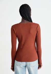 Vrouw met lang gevlochten haar staat met haar rug naar de camera, gekleed in een roestkleurig langemouwen top en lichtblauwe spijkerbroek tegen een witte achtergrond.