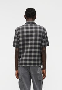 Πουκάμισο με κοντά μανίκια και καρό σχέδιο σε σκούρους τόνους μπλε και γκρι, με άνετη εφαρμογή, γιακά και λεπτομέρεια λογότυπου στο πίσω μέρος.