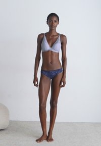 Lichtpaarse bralette van textuurstof met een kleine decoratieve knop, gecombineerd met donkerblauwe onderbroek met kant. Model staat op een beige tapijt.