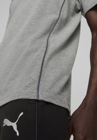 Grijs sport T-shirt met contrasterende donkergrijze stiksels, gecombineerd met zwarte broek met een wit Puma-logo. Focus op de stoftextuur.
