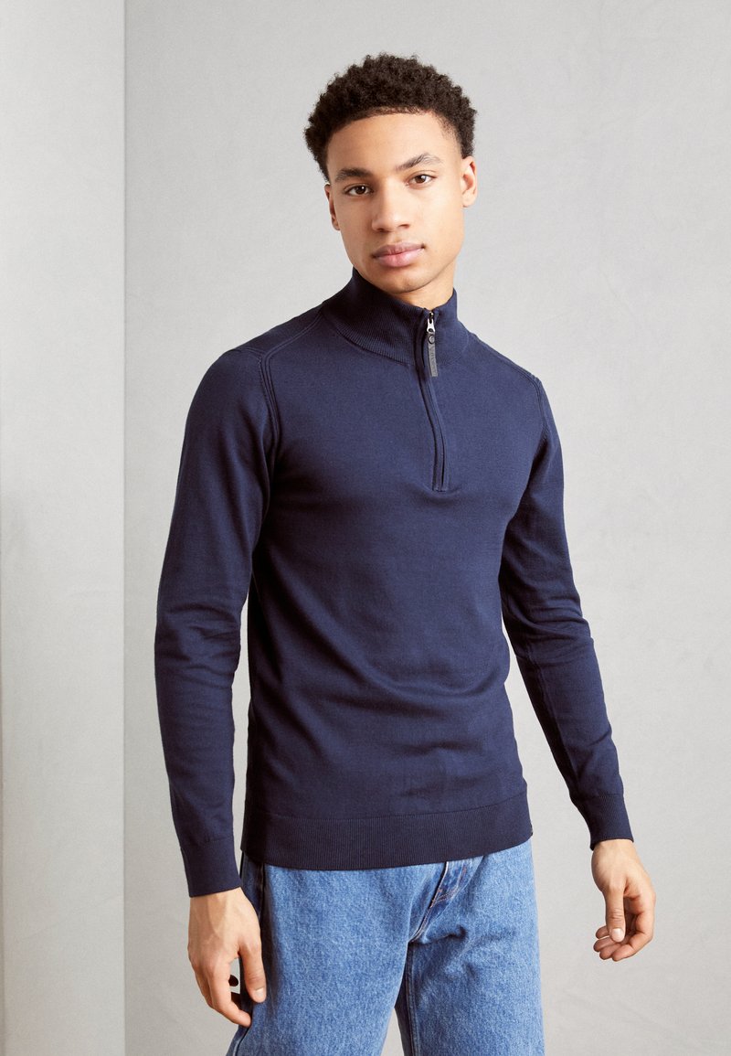 Suéter de punto azul marino con cremallera y cuello alto, mangas largas y puños acanalados, combinado con unos jeans de mezclilla azul claro.
