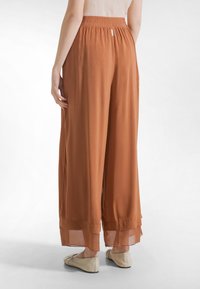 Løse bukser i rustfarve med en blød tekstur, udstyret med en elastisk, rynket talje og gennemsigtigt lagdelt kant ved anklerne.