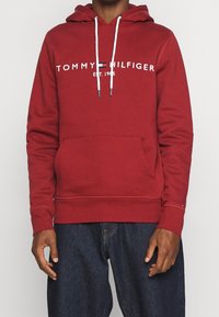Hombre con una sudadera roja de Tommy Hilfiger con cordones blancos y jeans azules, de pie contra un fondo claro y liso.